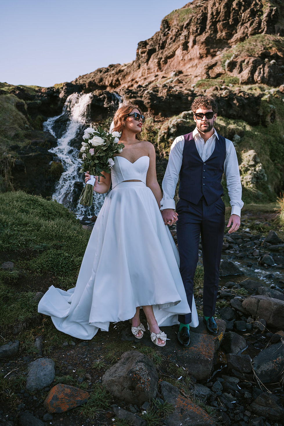 A couple beside a waterfall, the bride with a bouquet, the groom in a waistcoat. A gown by The Sposa Group with menswear by YSG Tailors, styling by Tamarra McNaught, founder of TITO STYLE, at Cairn Beach, Australia.