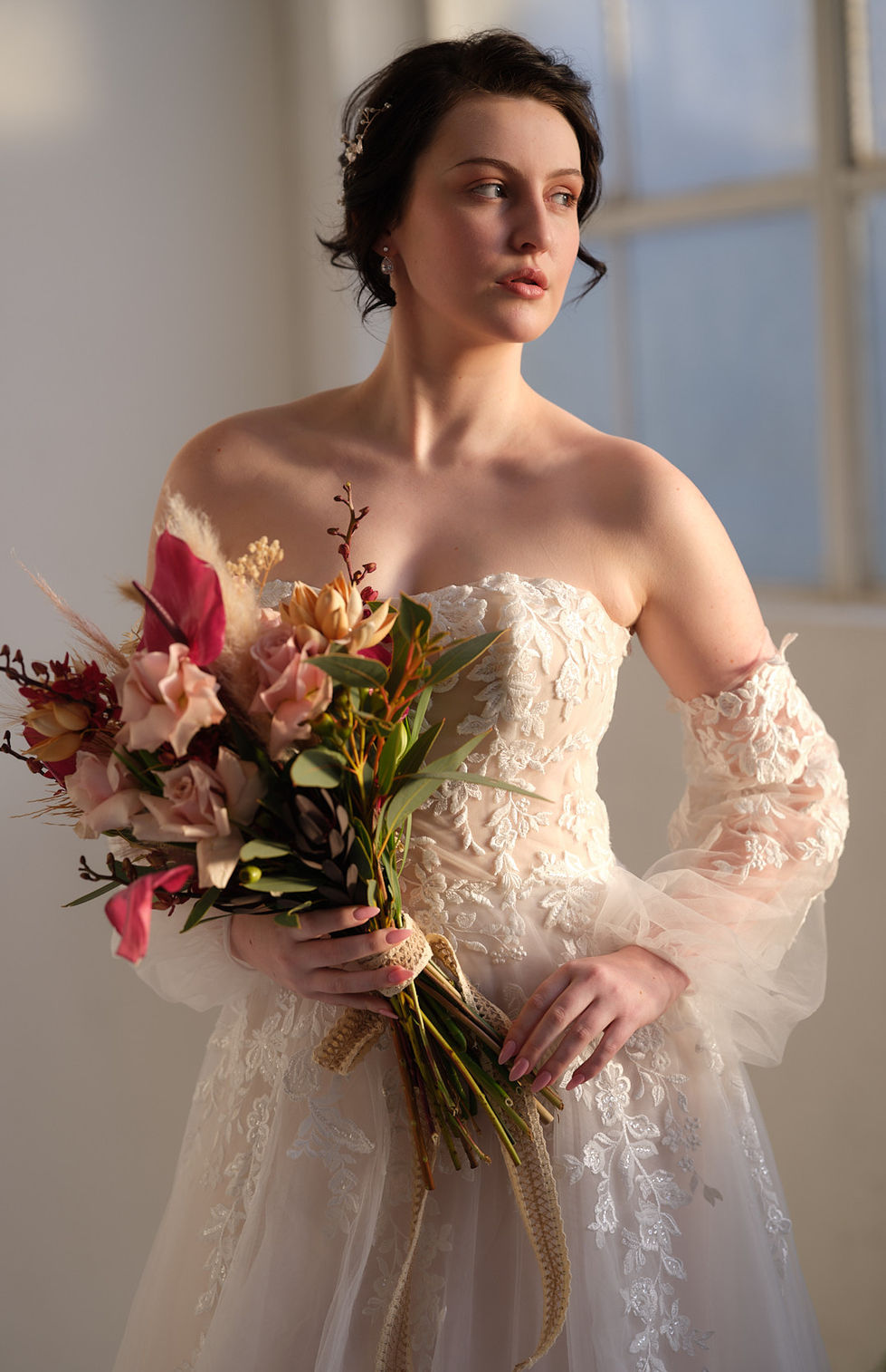 An off-shoulder lace gown with sheer sleeves, holding a soft pink and burgundy bouquet. A gown by The Sposa Group, with styling by Tamarra McNaught, founder of TITO STYLE, in Melbourne, Australia.