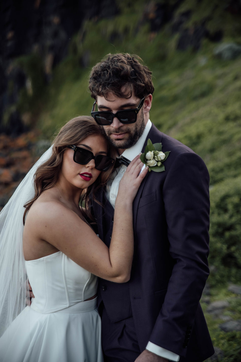 A couple in sunglasses, close and cool-toned, the groom in a dark suit. A gown by The Sposa Group with menswear by YSG Tailors and sunglasses by Gucci, styling by Tamarra McNaught, founder of TITO STYLE, at Cairn Beach, Australia.