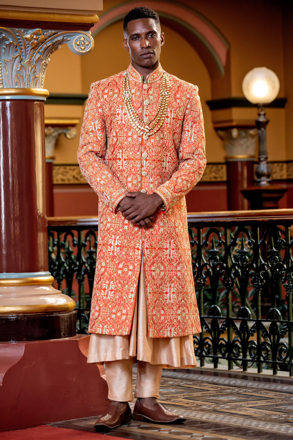 Male model in an orange and gold embroidered sherwani by Rajashree Bridal Studio on the balcony of The Hotel Windsor, styled by TITO STYLE.