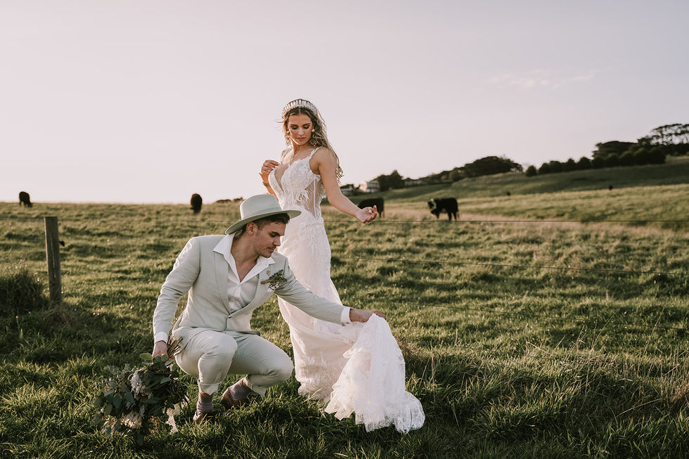 The groom kneeling to arrange the bride's train in a field. A gown by The Sposa Group with menswear by YSG Tailors, styling by Tamarra McNaught, founder of TITO STYLE, at Cairn Beach, Australia.
