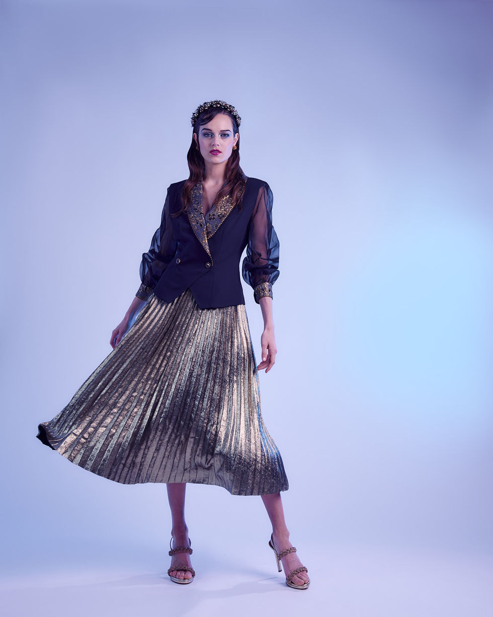 A shimmering gold pleated skirt and black jacket finished with a jewelled crown, full length in a blue-lit studio in Melbourne, Australia, featuring pieces by Anthea Crawford, Dolci Firme and We Are The Others, styling by Tamarra McNaught, founder of TITO STYLE.