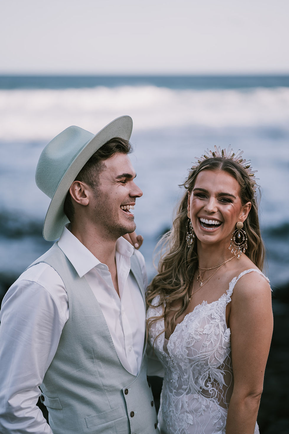 A couple close together by the sea, the bride in a floral crown, the groom in an olive suit and hat. A gown by The Sposa Group with menswear by YSG Tailors and a hat by Lack of Color, styling by Tamarra McNaught, founder of TITO STYLE, at Cairn Beach, Australia.