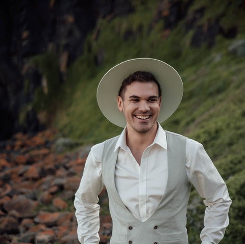 Groom in light linen suit and wide-brim hat smiling confidently, standing on rocky coastal path before wedding ceremony