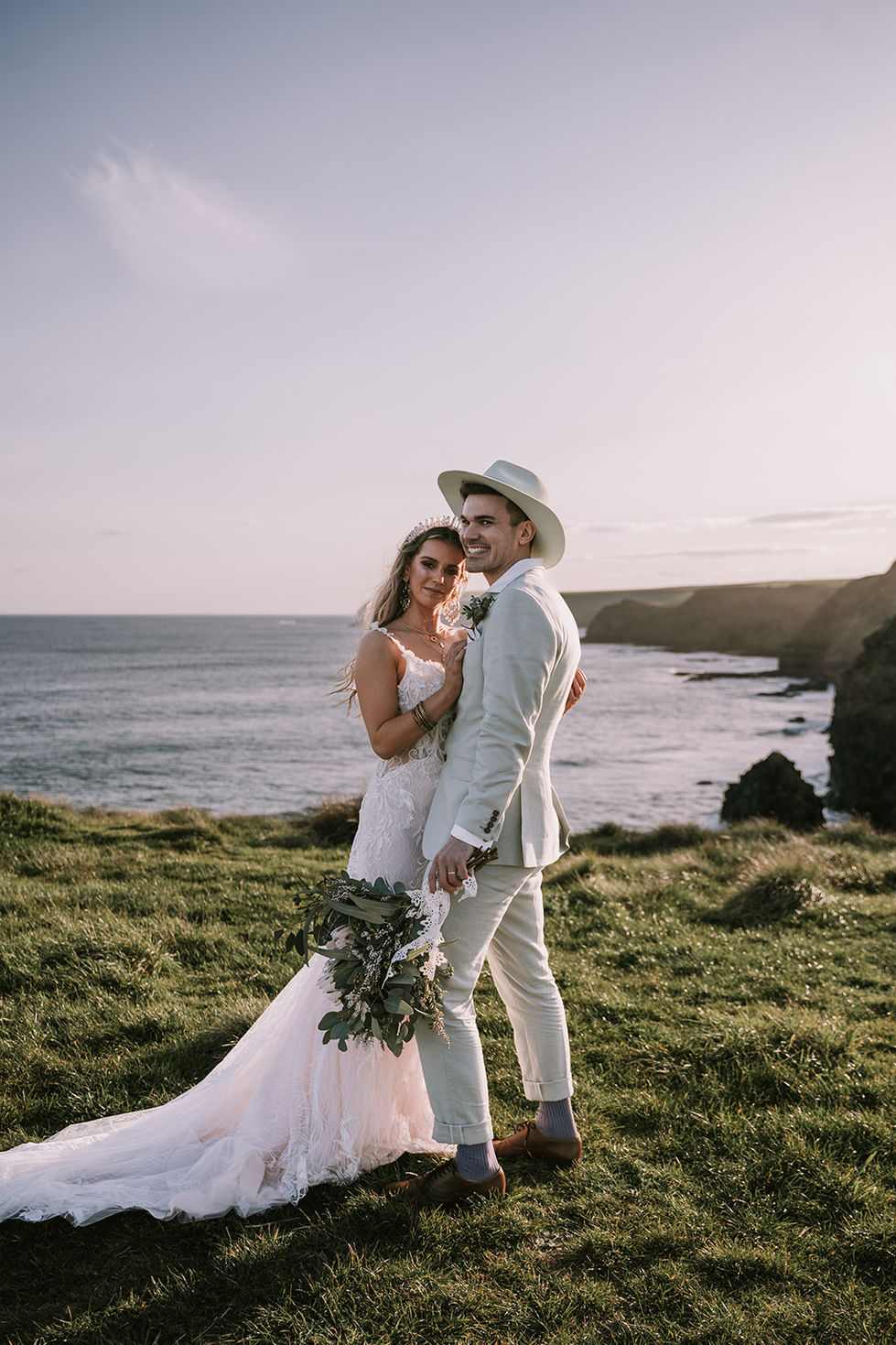A couple on the coastal cliffs, the bride holding a bouquet, the groom in an olive suit and hat. A gown by The Sposa Group with menswear by YSG Tailors and a hat by Lack of Color, styling by Tamarra McNaught, founder of TITO STYLE, at Cairn Beach, Australia.
