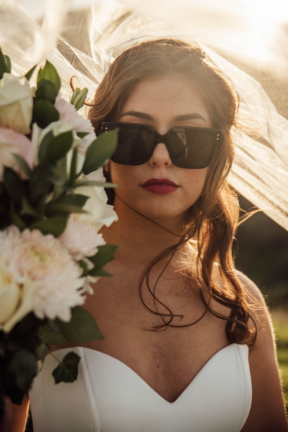 A bride in sunglasses holding white lilies, backlit by the sun. A gown by The Sposa Group with sunglasses by Gucci, styling by Tamarra McNaught, founder of TITO STYLE, at Cairn Beach, Australia.