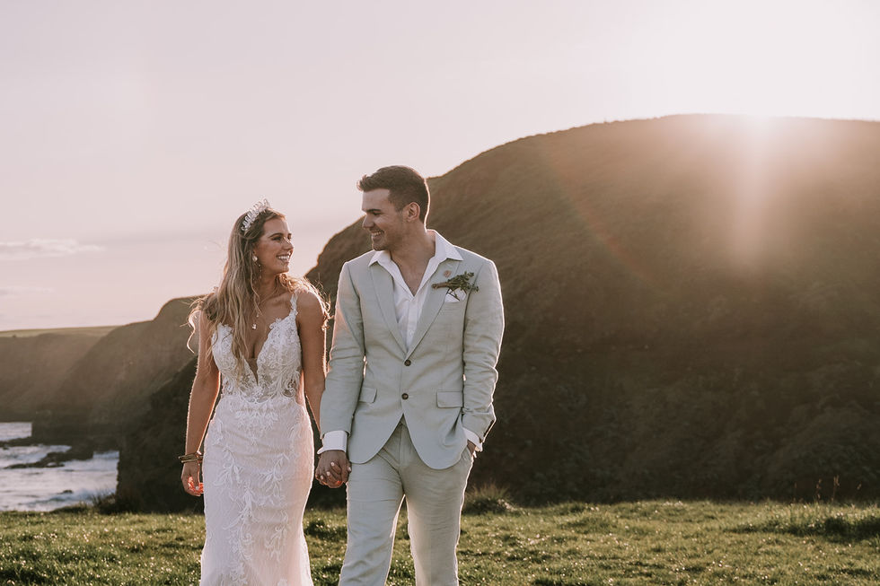 A couple walking the headland in golden light, the bride in a lace mermaid gown, the groom in olive tailoring. A gown by The Sposa Group with menswear by YSG Tailors, styling by Tamarra McNaught, founder of TITO STYLE, at Cairn Beach, Australia.