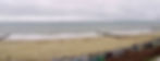 Cloudy beach scene with calm waves, sandy shore, colorful beach huts, and a lone person walking. Overcast sky sets a serene mood.