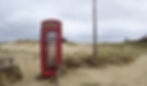 Red phone box on sandy dunes with grass, under cloudy skies. A wooden post stands nearby. The scene is isolated and tranquil.