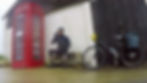 Cyclist with helmet sits by red phone booth, drinking from a bottle. Bicycle leans against wall with panniers. Overcast, outdoor setting.