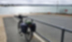 Bicycle with panniers near a calm sea, cloudy sky. Ferry and "No Fishing" sign in background. Peaceful coastal scene.