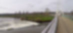Modern bridge over a river with flowing water; green fields and bare trees in the background under a cloudy sky.