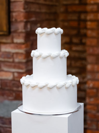 A three-tier all-white wedding cake from The Piped collection, featuring repetitive horizontal shell piping details on each tier, displayed against a rustic brick wall at 26 Bridge in Brooklyn.