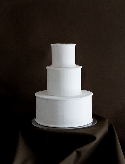 A three-tier all-white minimalist wedding cake from The Border collection, featuring clean architectural lines and precise border detailing, set against a dark, moody background.