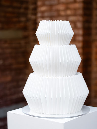 A three-tier all-white modern wedding cake from The Origami collection, featuring hand-cut geometric pleats and sharp architectural lines, displayed at a Brooklyn industrial venue.