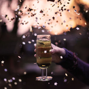 A hand holds a champagne glass amidst falling star-shaped confetti at sunset. The scene is festive and warm with a blurred background.