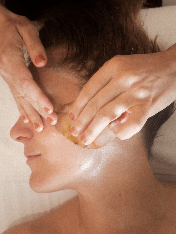 Person receiving a gua sha facial massage, lying down in a calm setting. The mood is serene and relaxed.