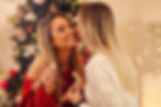 Two women smiling, one in a red sweater, sharing a sweet moment by a decorated Christmas tree with warm lights in the background.