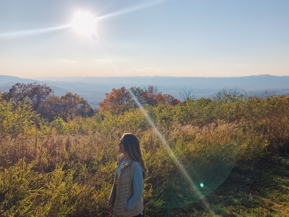 A girl standing in front of a mountain overlook with a sunburst in the sky.