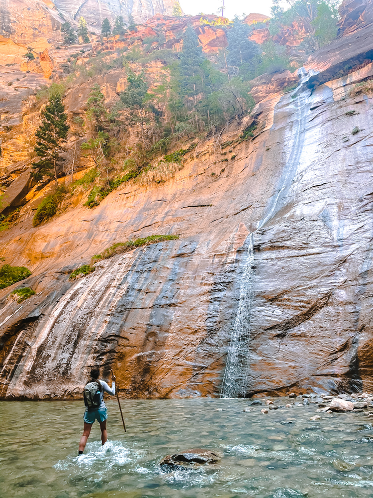 A woman is standing in the Virgin River next to a waterfall on the Narrows hike