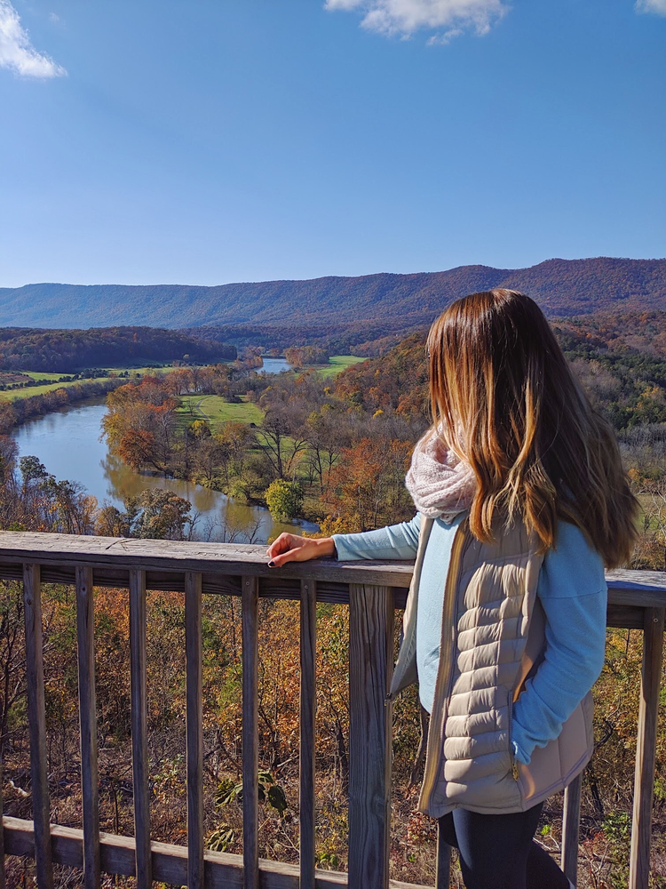A girl stands against a railing overlooking a river that winds through the mountains. Fall color