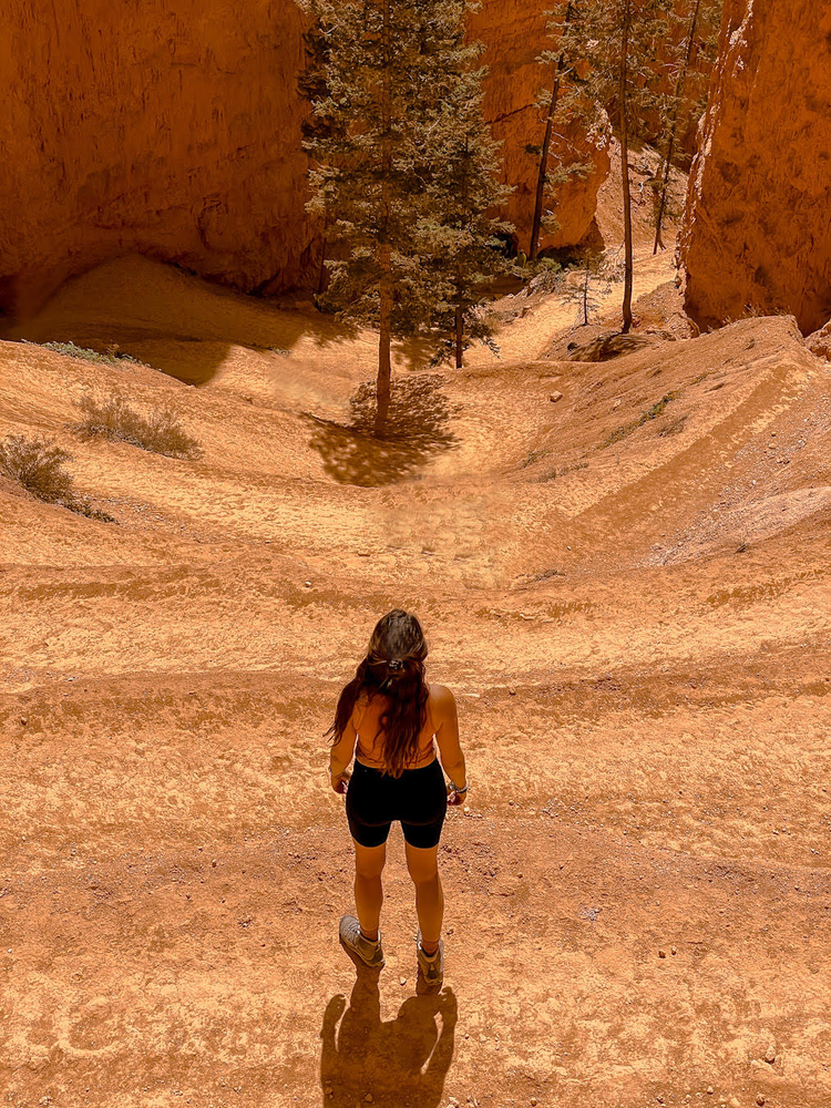 A woman stands on a sandy trail overlooking a series of switchbacks 