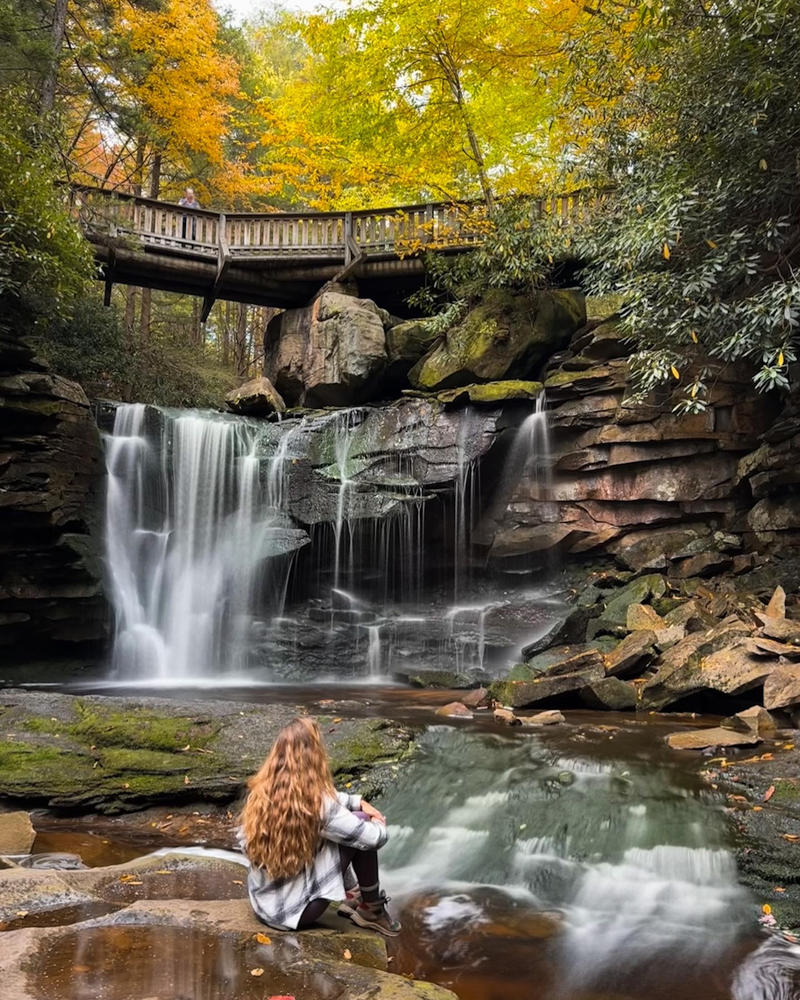 A girl sits looking at a waterfall with a bridge above it