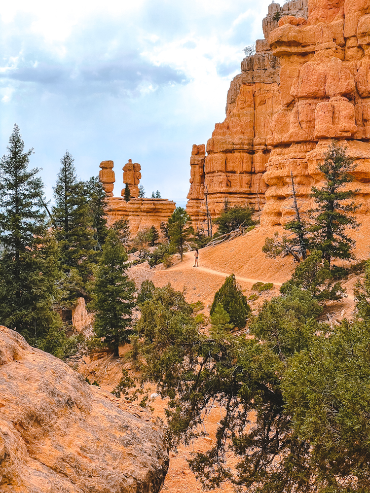 A woman can be seen in the distance standing on a trail among red rock formations