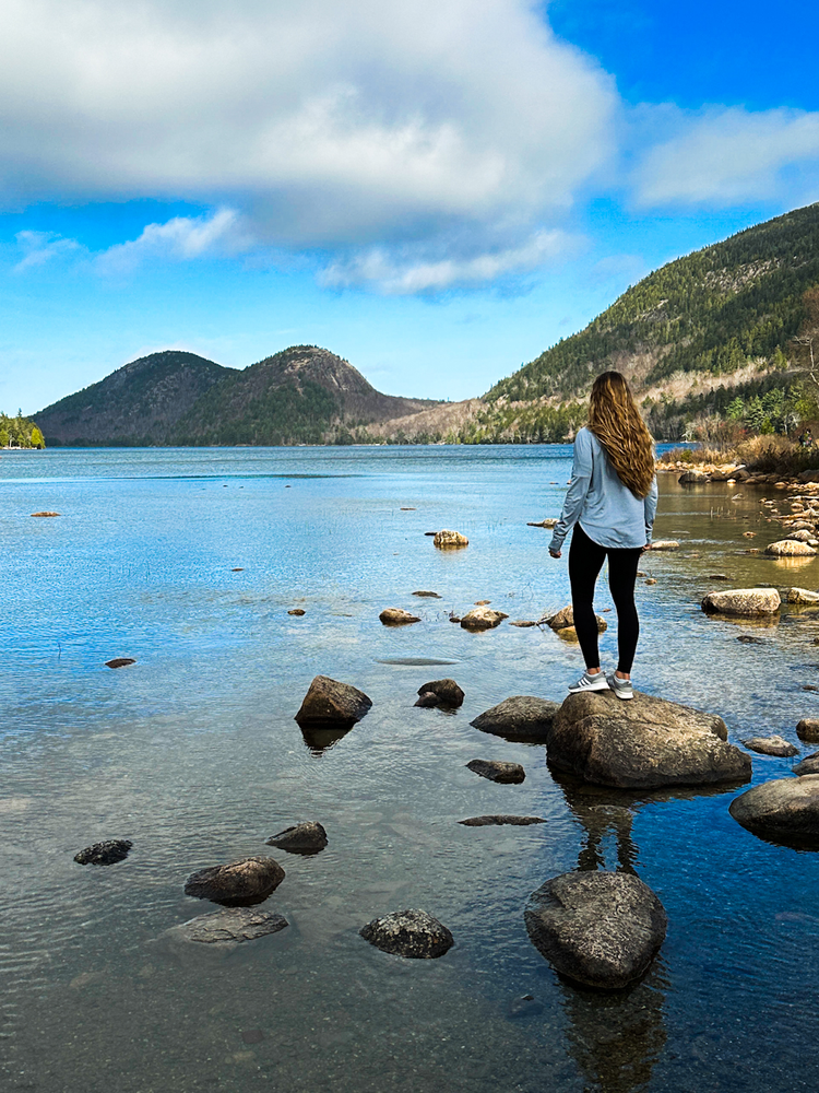 A girl stands on a boulder in a pond looking out at the mountains