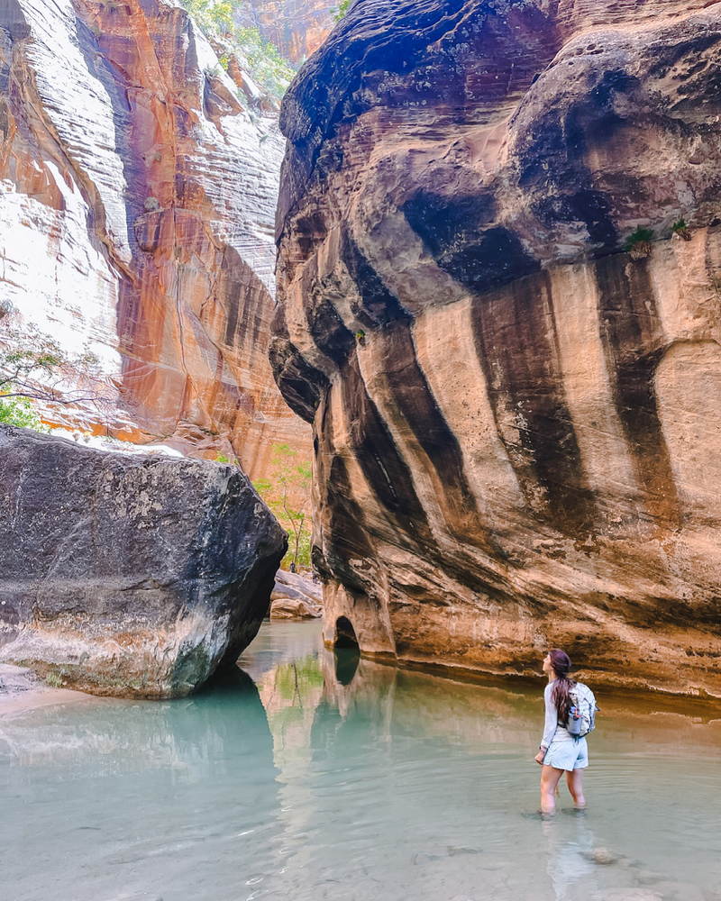 A woman standing in a milky blue river amongst towering rock formations with unique striations