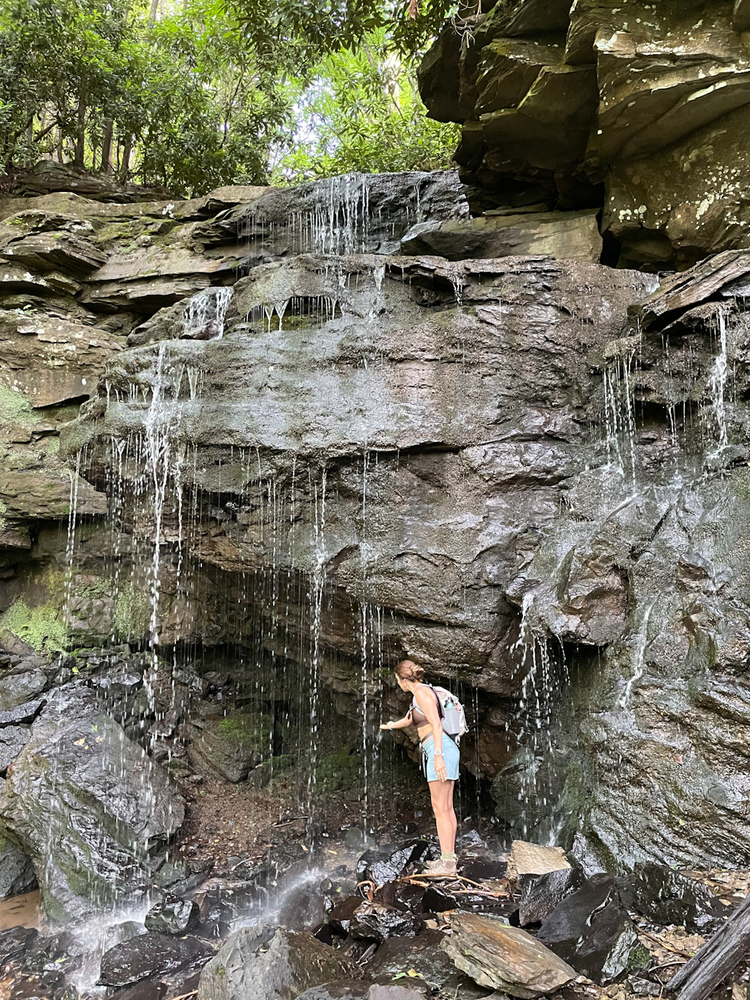 A girl is standing behind a waterfall reaching out to touch the water