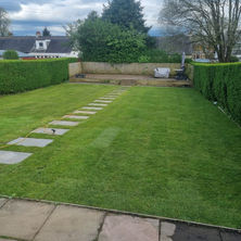 A well-maintained green lawn features stone stepping stones leading to a hedge-lined garden