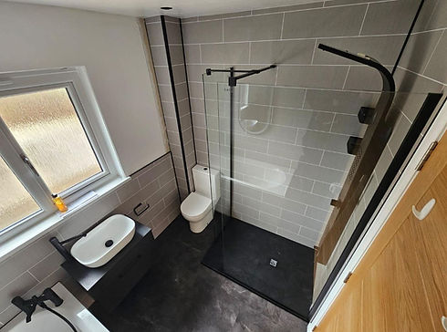Modern bathroom with a sleek design featuring gray tiled walls, a glass shower enclosure, white sink, toilet, and a window