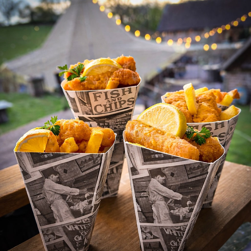 Four paper cones with fish and chips, garnished with lemon and parsley, are arranged outdoors. Warm, festive lights in the background.