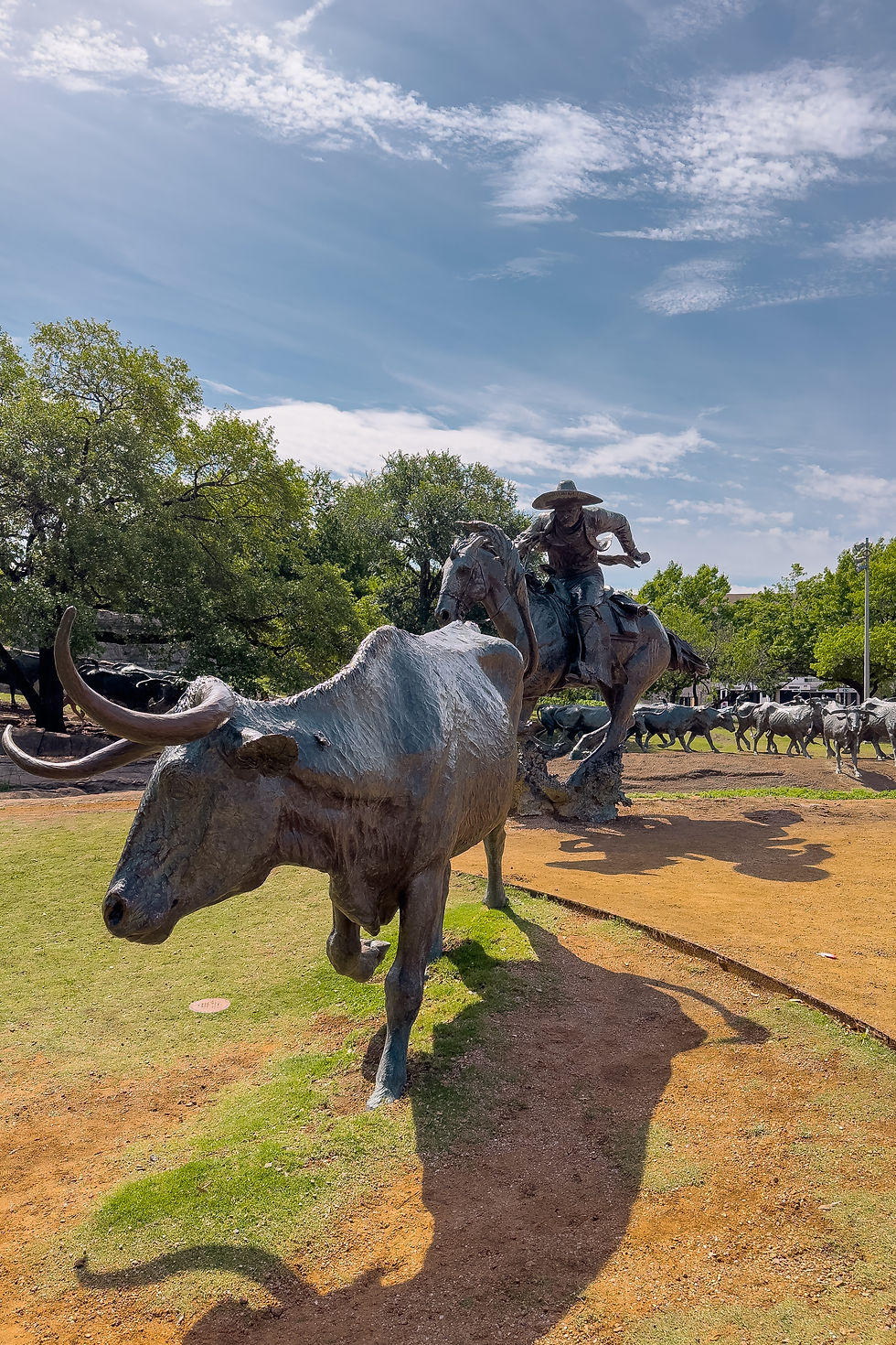 Statues de cowboy et taureau en bronze sur un chemin terreux, ciel bleu au-dessus. Ambiance dynamique et historique.