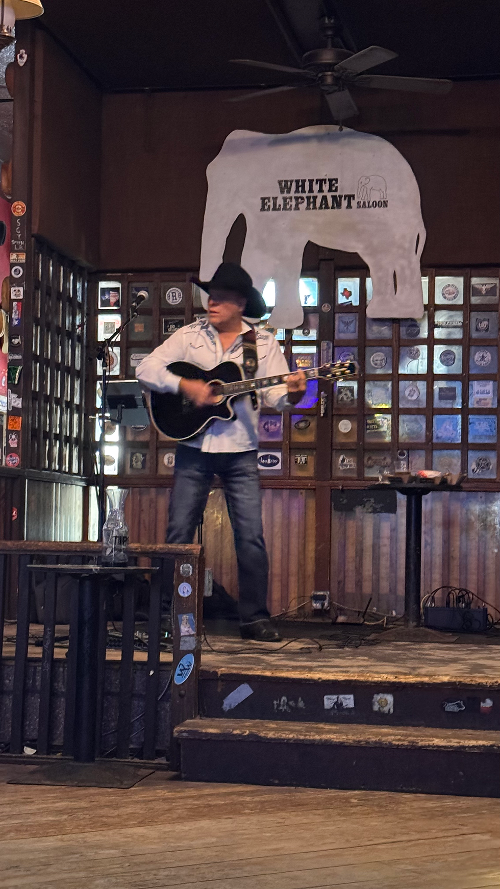 Un musicien en chemise blanche et chapeau noir joue de la guitare sur scène. Fond de saloon avec une enseigne "White Elephant". Ambiance chaleureuse.