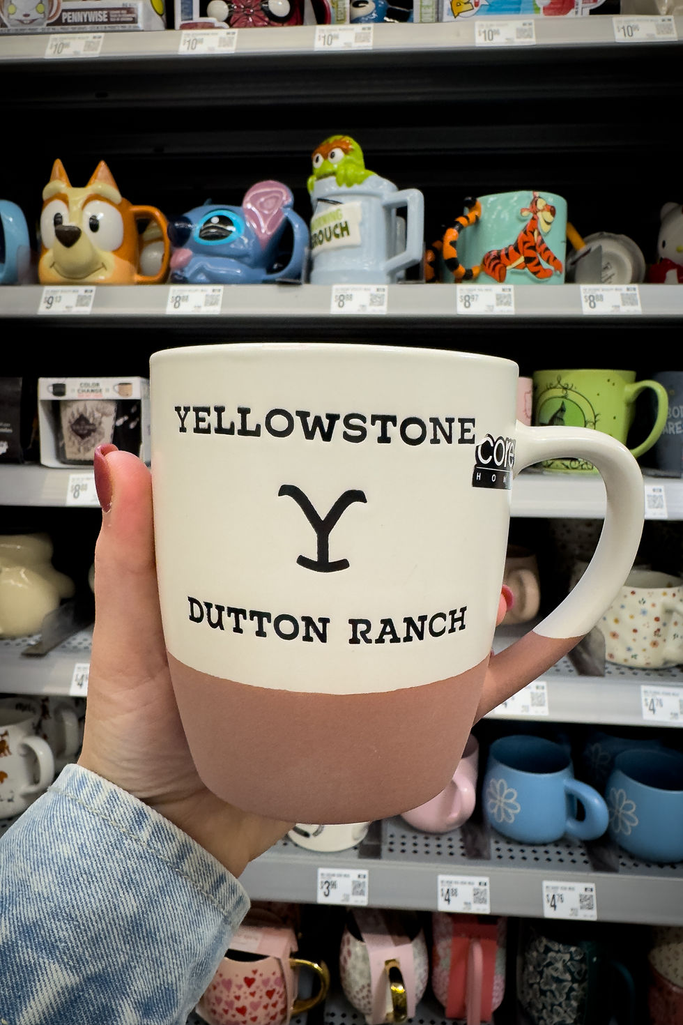 Tasse blanche à motif rose, texte "YELLOWSTONE DUTTON RANCH", tenue par une main en chemise bleue, étagères de mugs colorés en fond.