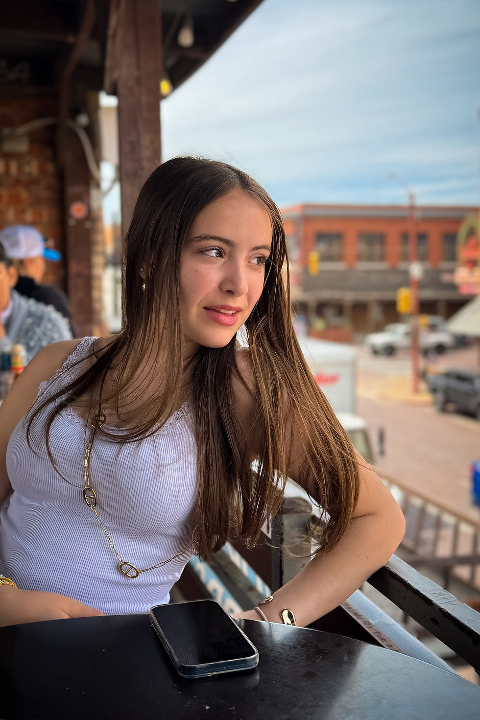 Jeune femme assise en terrasse, regardant au loin, portant un top blanc. Un smartphone est posé sur la table. Fond urbain ensoleillé.