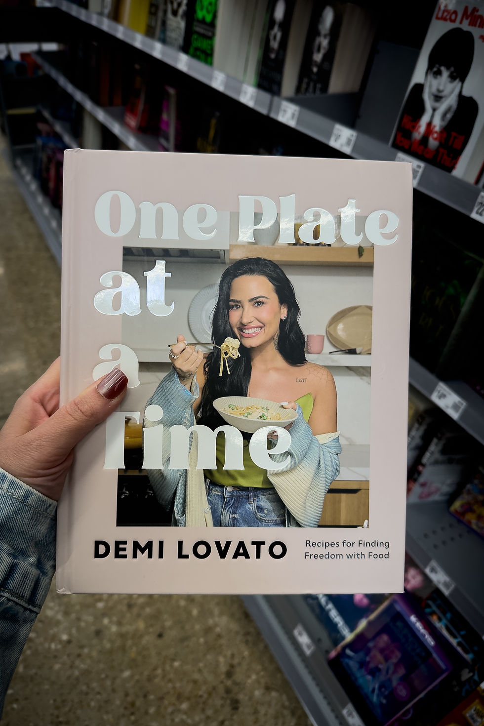 Une main tient le livre "One Plate at a Time" de Demi Lovato. Elle sourit avec un bol de pâtes. Rayon de livres en fond.