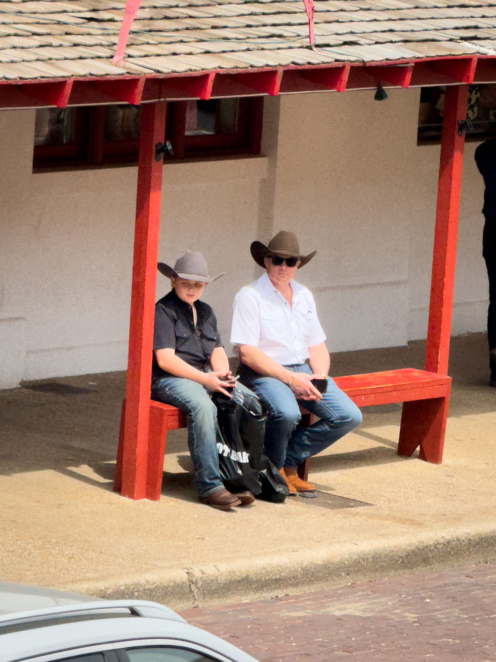 Deux personnes en chapeaux de cowboy assises sur un banc rouge, l'air détendu. Décor urbain avec toit en bois et sac noir.