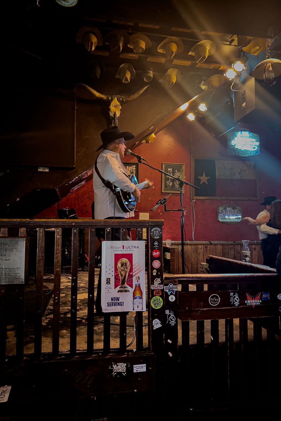 Un musicien en chemise blanche et chapeau joue de la guitare sur scène dans un bar au décor western, avec lumières tamisées et affiche.