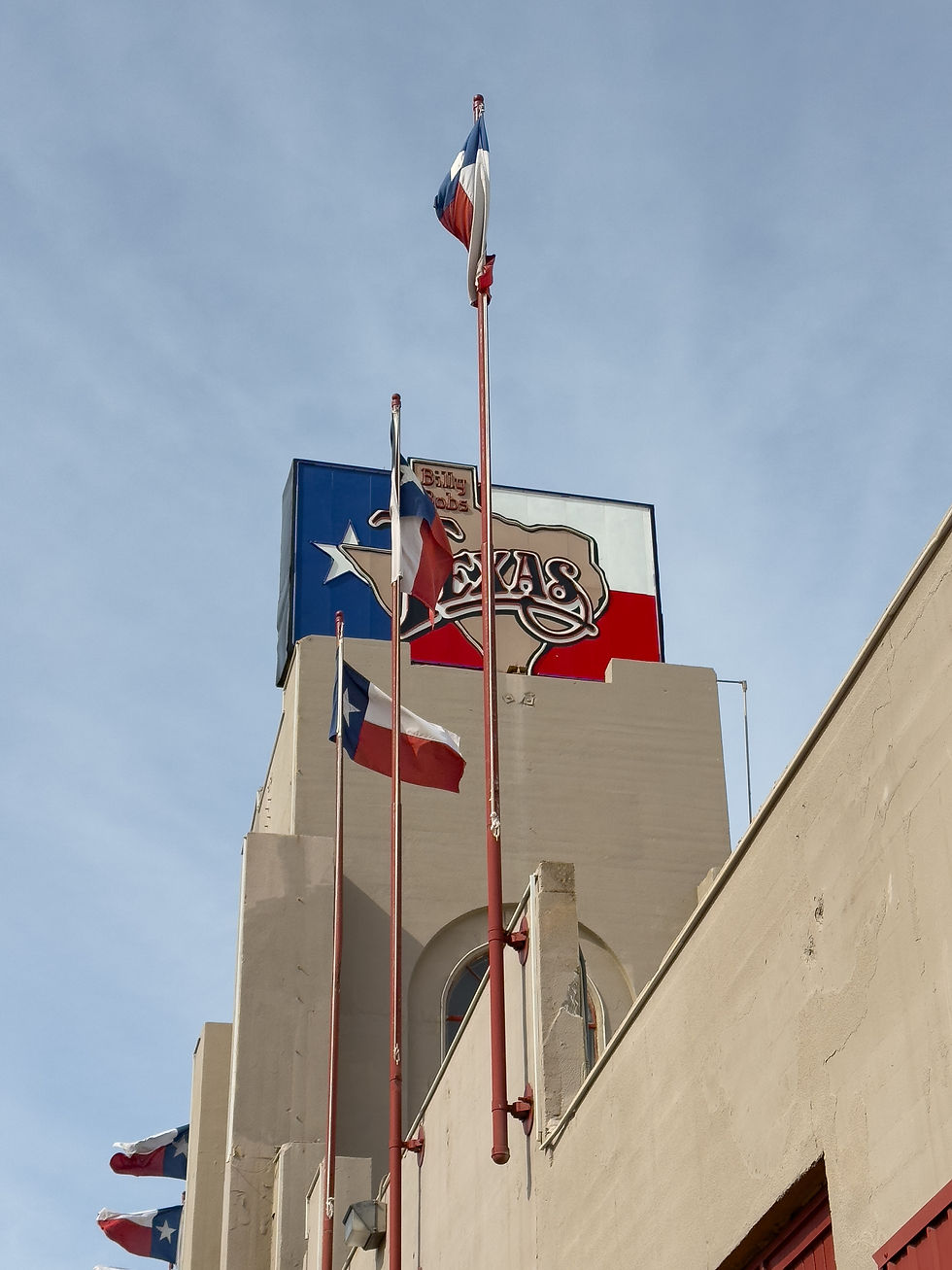 Trois drapeaux texans flottent sur un bâtiment beige. Une enseigne bleue et rouge indique "Texas". Ciel clair en arrière-plan.