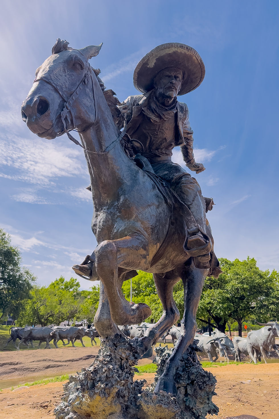 Statue en bronze d'un cowboy sur un cheval galopant, devant un troupeau. Ciel bleu, arbres verts en arrière-plan, ambiance western.