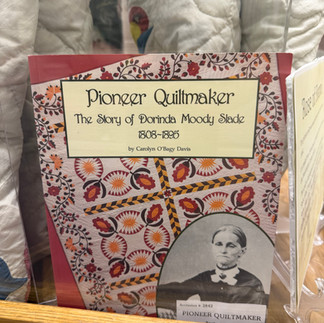 A display case featuring the book “Pioneer Quiltmaker: The Story of Dorinda Moody Slade” next to the historic quilt she created.