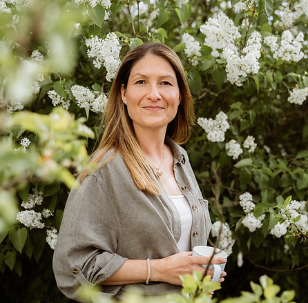 Frau steht entspannt vor blühenden Sträuchern mit einer Tasse in der Hand – symbolisiert Ruhe, Achtsamkeit und naturnahes Coa