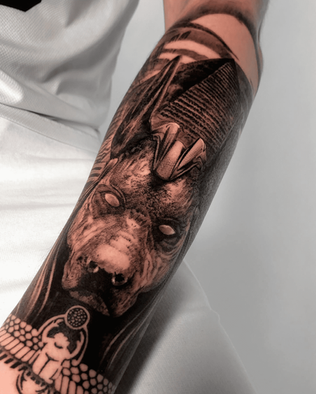 tattoo of the statue of the Egyptian god Anubis on the forearm in Black and White realism by APZ in Copenhagen, Denmark.In Copenhagen, APZ is a skilled tattoo artist trained by some of the best tattoo artists in the world. He specializes in classic realism tattoos using a modern black and white style, focusing on connected designs and full sleeve tattoos that enhance his clients’ style. His sculpture Anubis tattoo on forearm speaks volumes with lifelike realistic details.