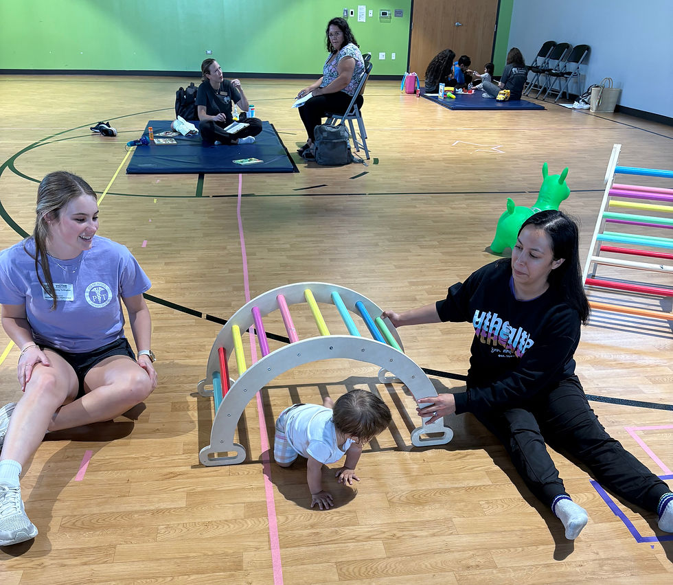 Two ACN student clinicians support an infant's play during a mini-screening event while her mother looks on in the background, consulting with a clinician.