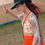 Thumbnail: Woman wearing hat and orange pants holding a drink, smiling widely.