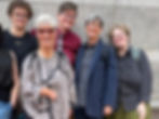 A group of five people posing as a group and smiling. From left to right, a white-skinned person with short curly brown hair and glasses; a white-skinned person with short gray hair and glasses in a gray top; a white non-binary person with short brown hair and glasses; a white-skinned person with short gray hair and glasses in a blue blazer; a white-skinned person with short curly blonde hair and glasses.