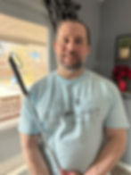 A white-skinned, masc-presenting person with short gray hair and a gray beard standing in front of a window, holding what a appears to be a white cane, wearing a baby blue disabled anthropologist t-shirt. 